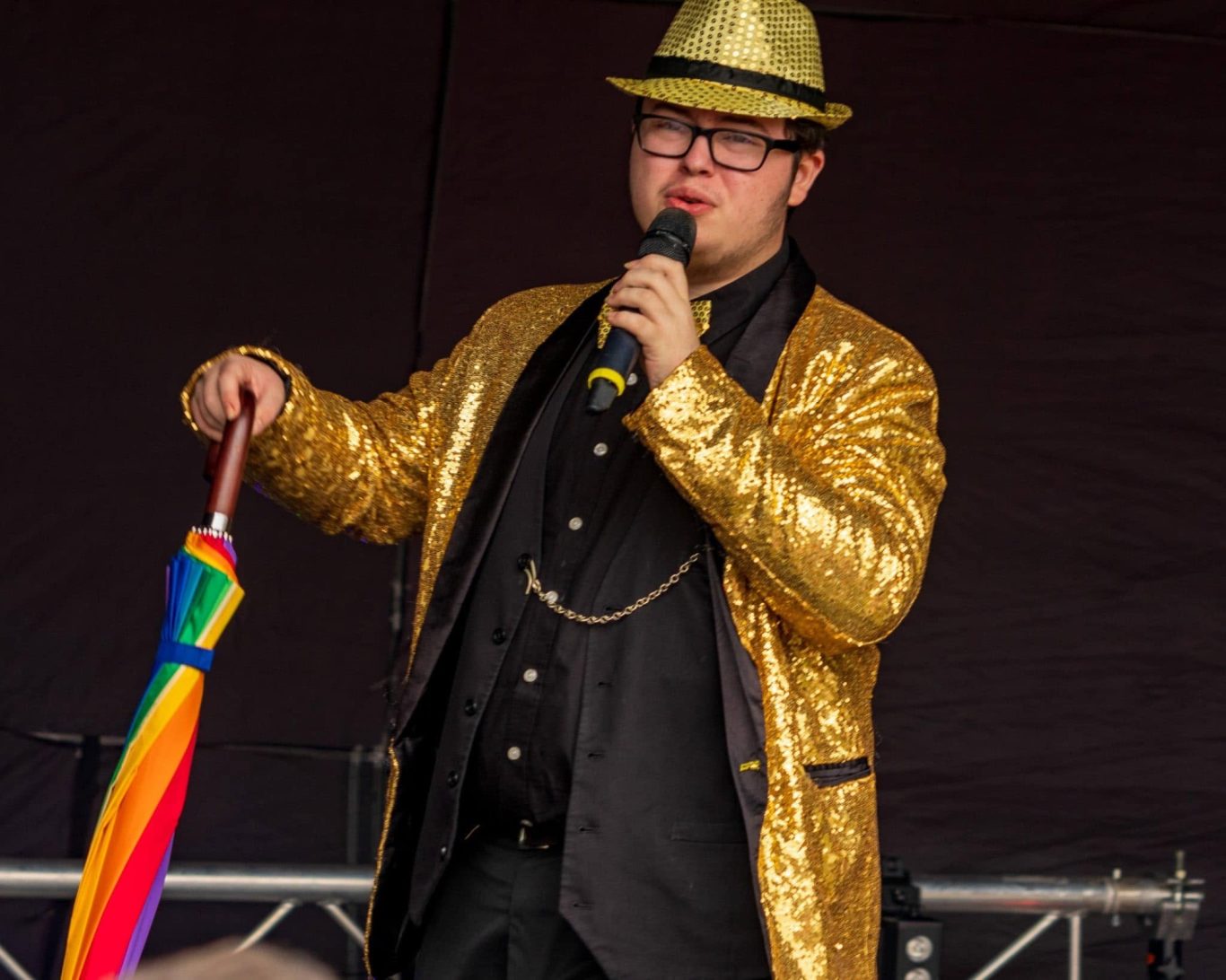 GYG - 2024 Person in a gold sequined suit holding a rainbow umbrella while speaking into a microphone.