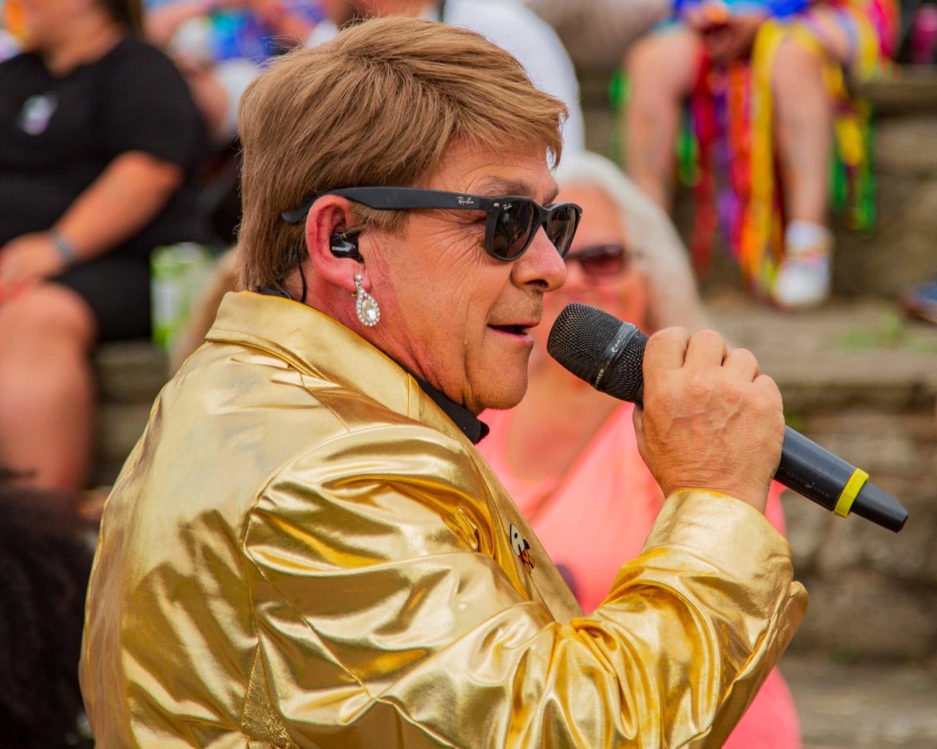 Elton John Tribute A performer in a shiny gold jacket and sunglasses singing into a microphone.