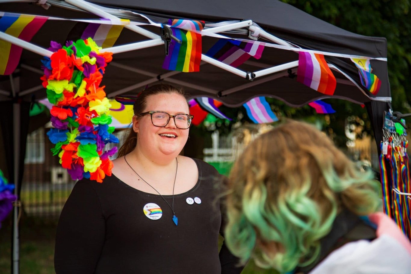 A smiling person at a festival booth, surrounded by rainbow decorations.