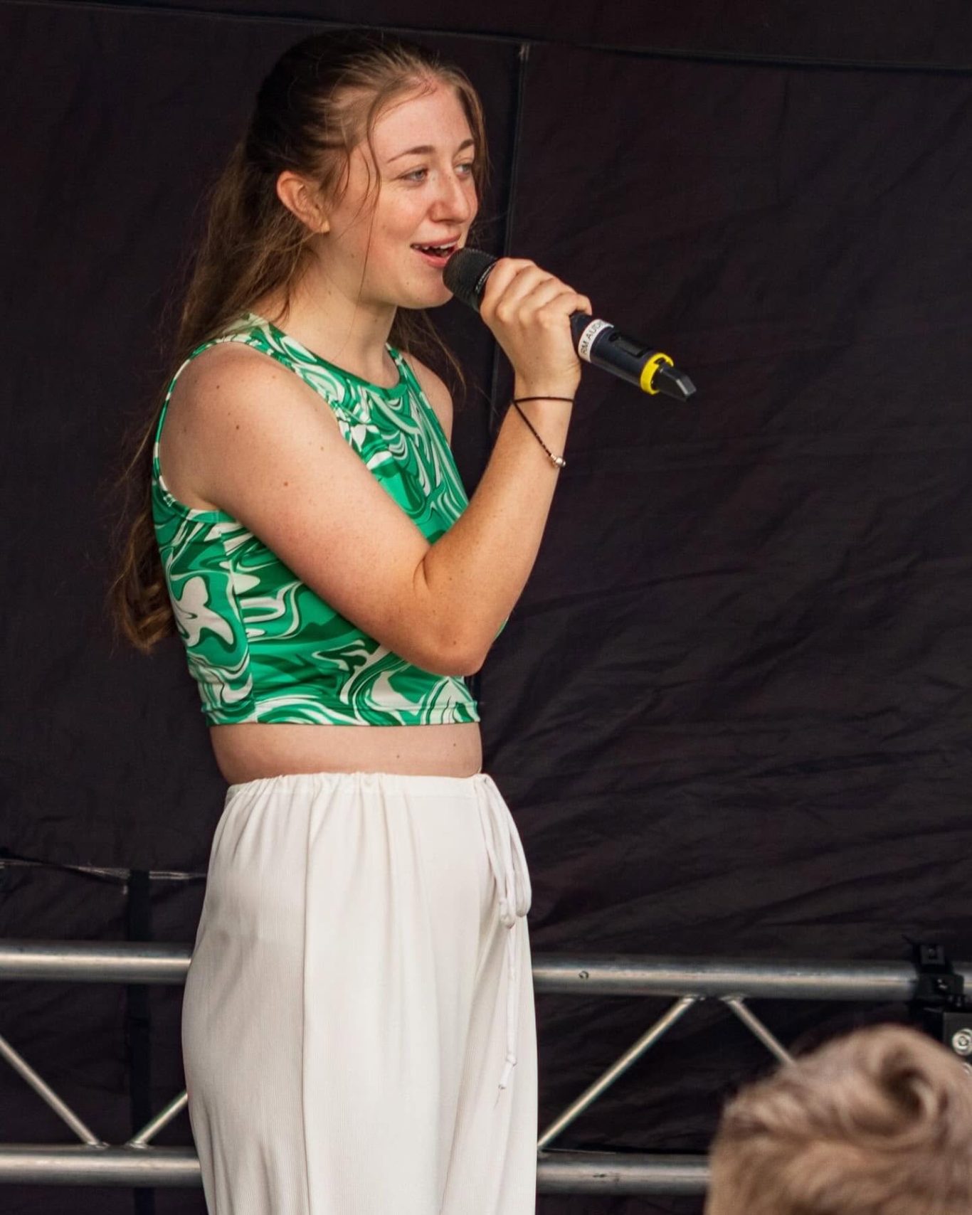 GYG - 2024 A young woman sings passionately on stage, wearing a green crop top and white trousers.