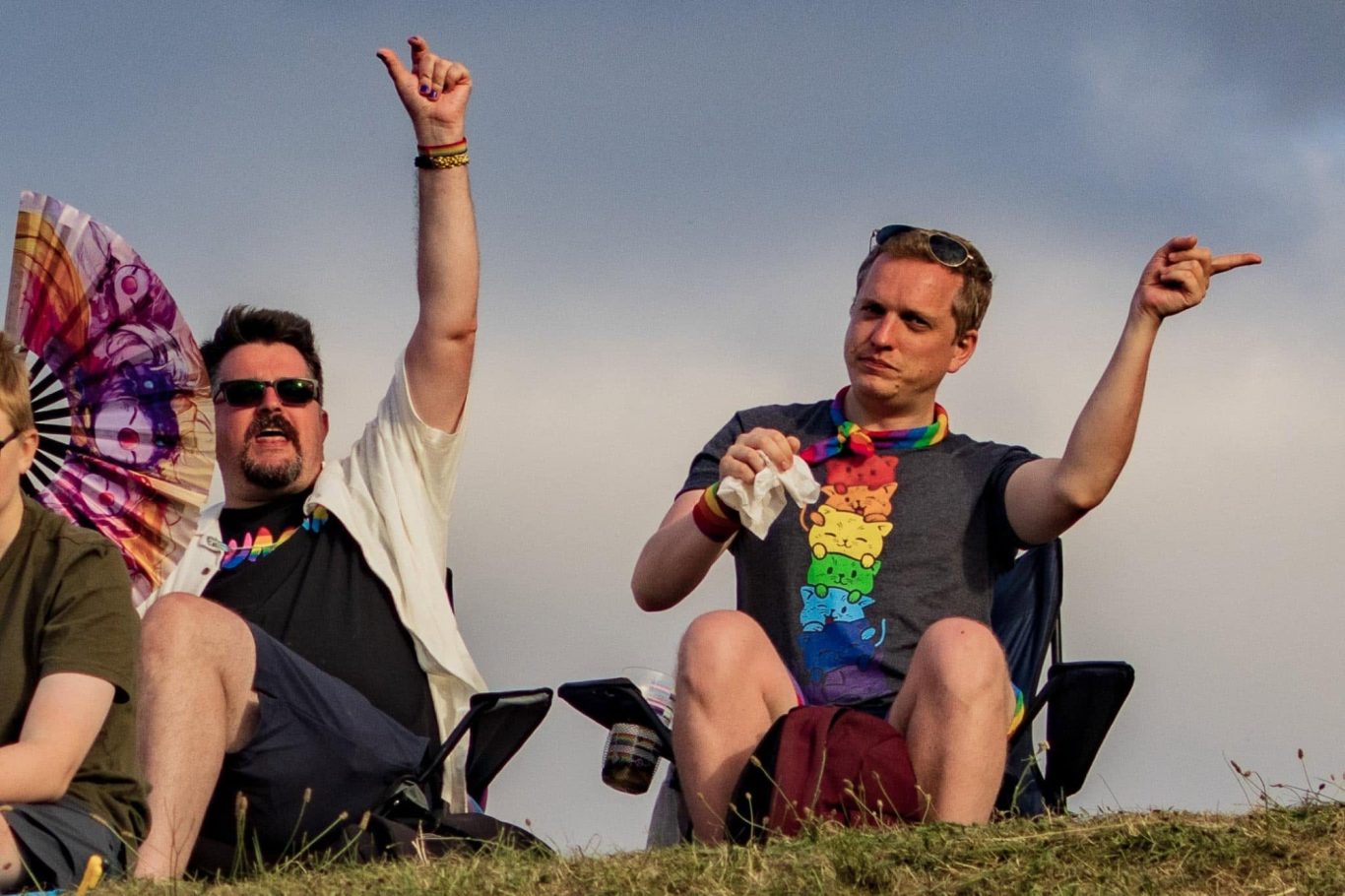 Two men sit on a hillside, wearing rainbow attire and celebrating with cheerful gestures.