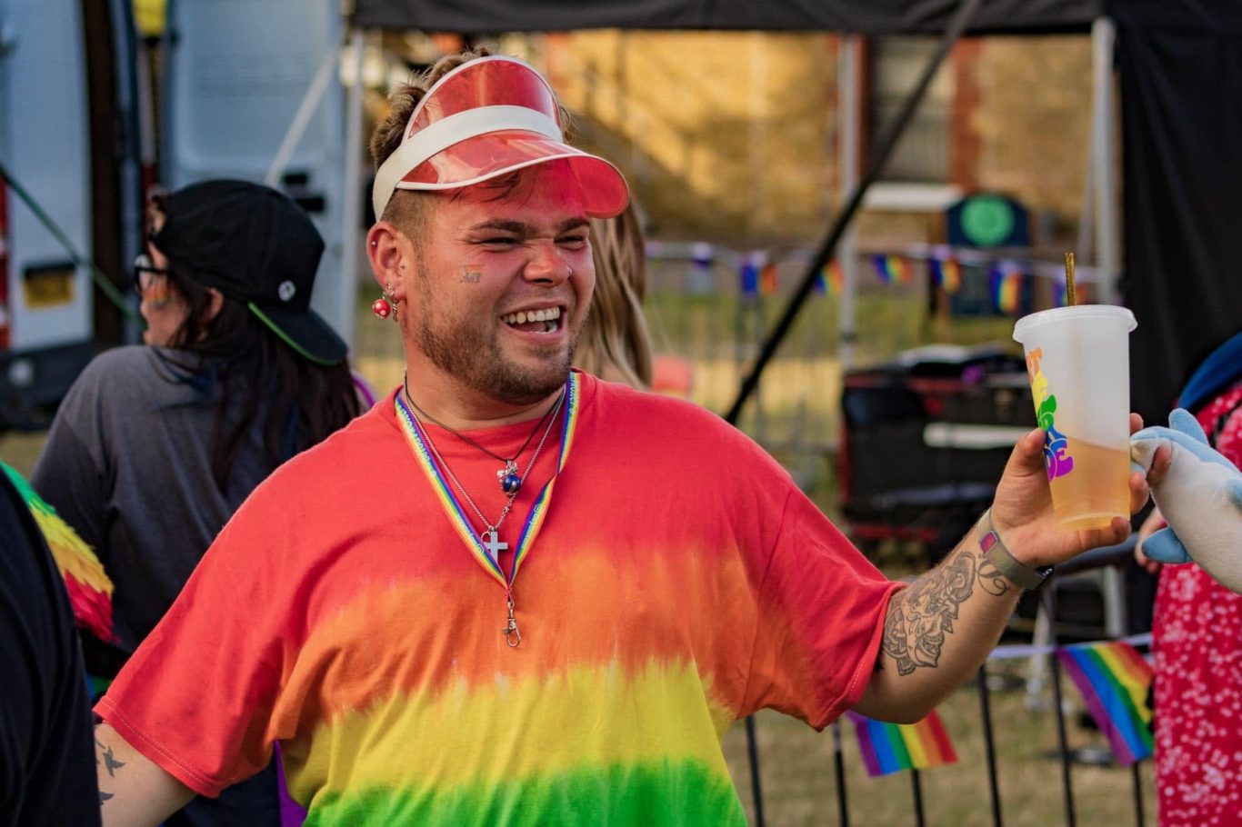 Person in a rainbow tie-dye shirt holds a drink, smiling at a festive outdoor event.