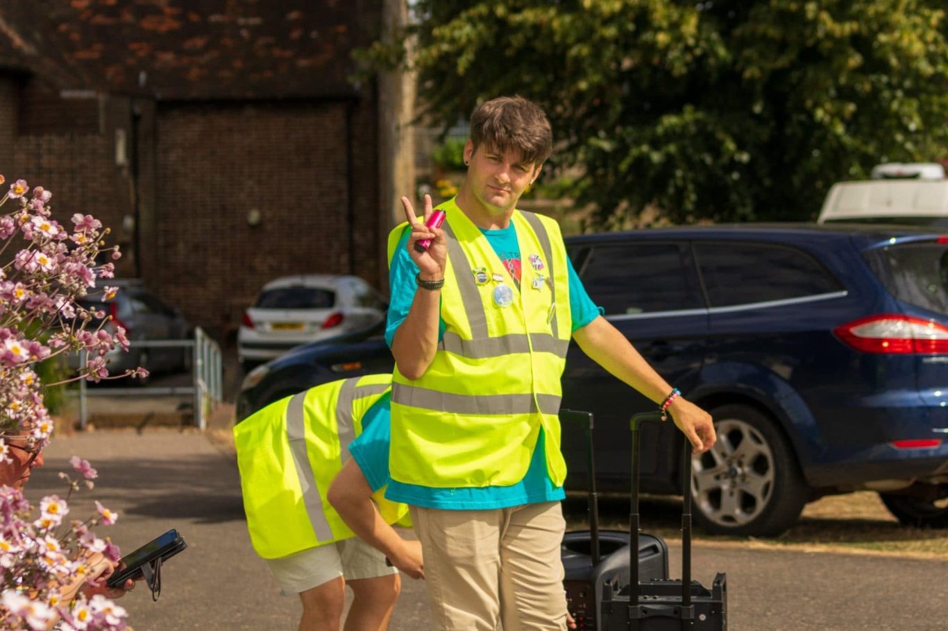 A young person in a yellow vest holds up a peace sign while others work nearby.