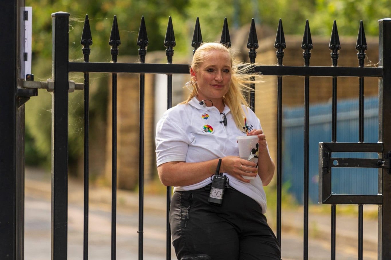 A woman with long blonde hair leans against a black fence, holding a device.