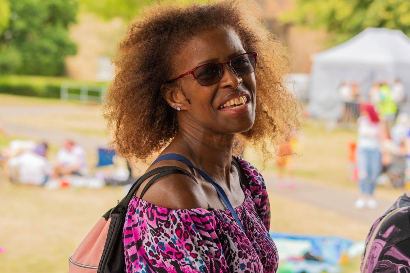 Smiling woman with sunglasses and curly hair, wearing a floral top in a park setting.