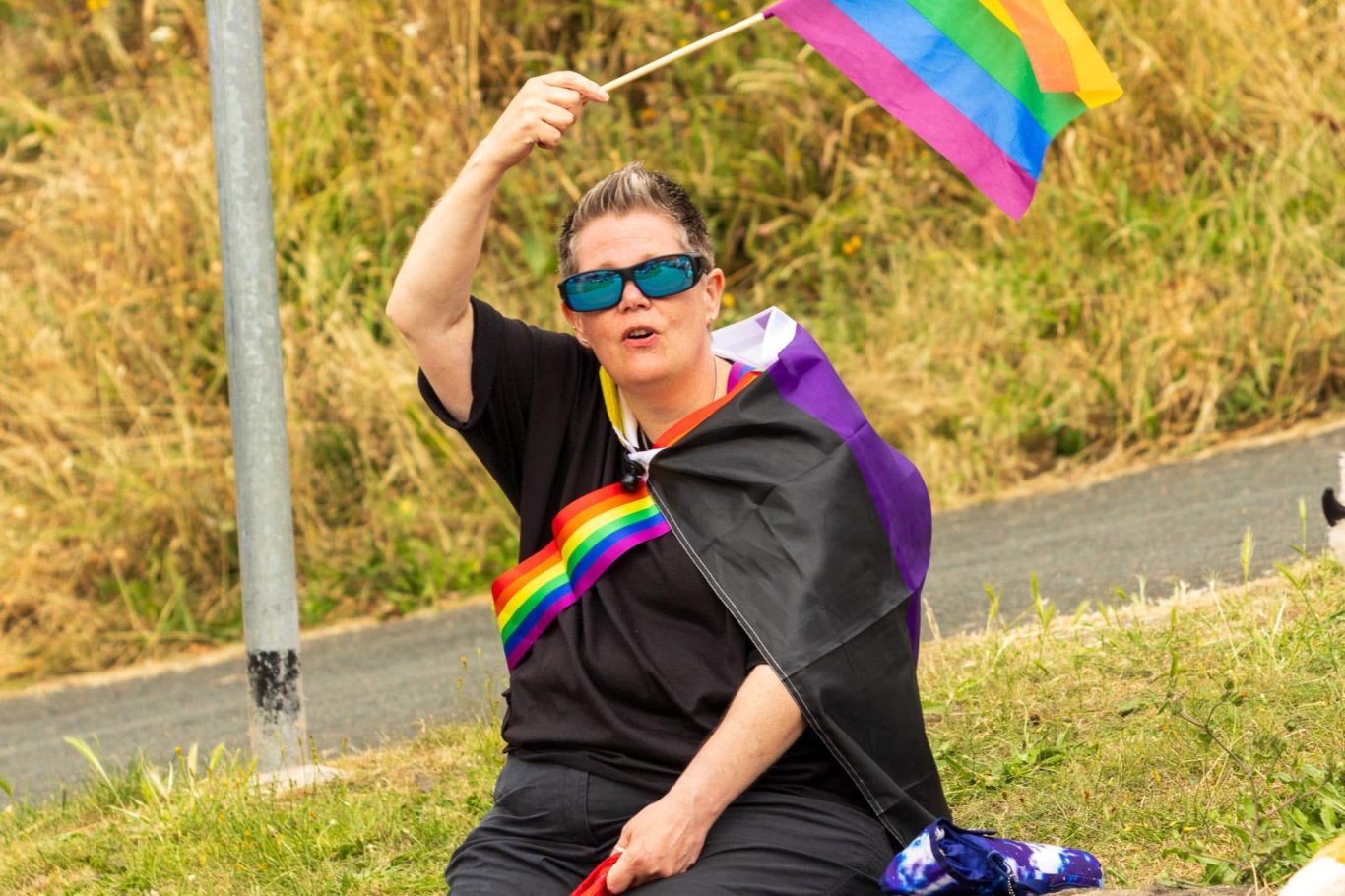A person wearing a rainbow flag and sunglasses waves a pride flag while sitting on grass.
