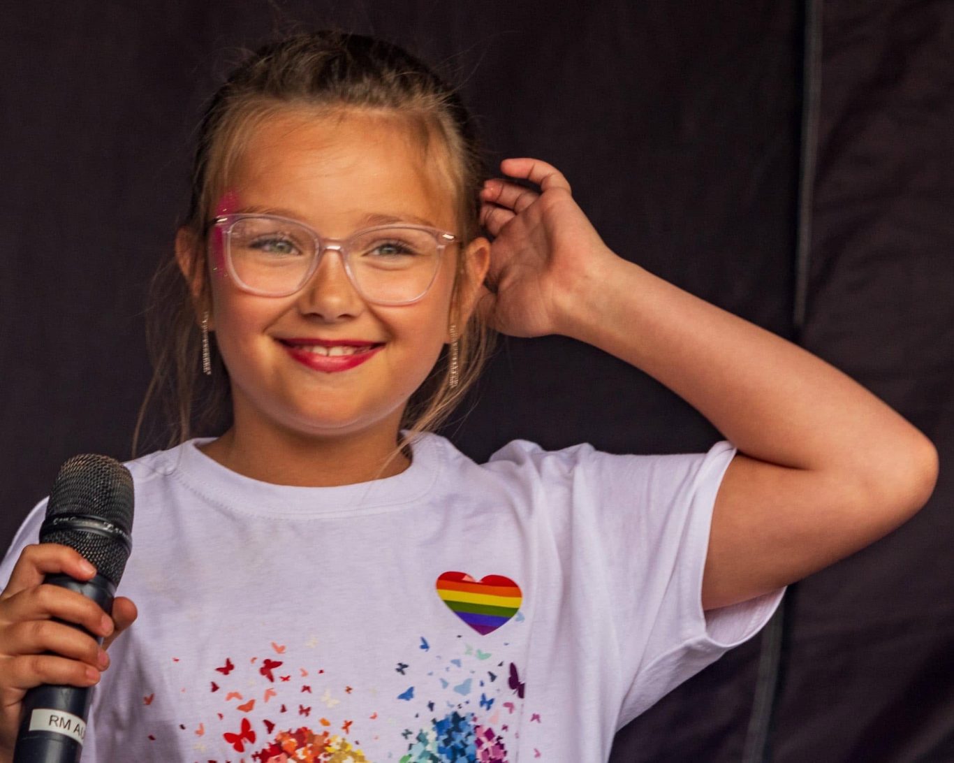 GYG - 2024 Smiling girl wearing glasses and a rainbow heart t-shirt, holding a microphone.