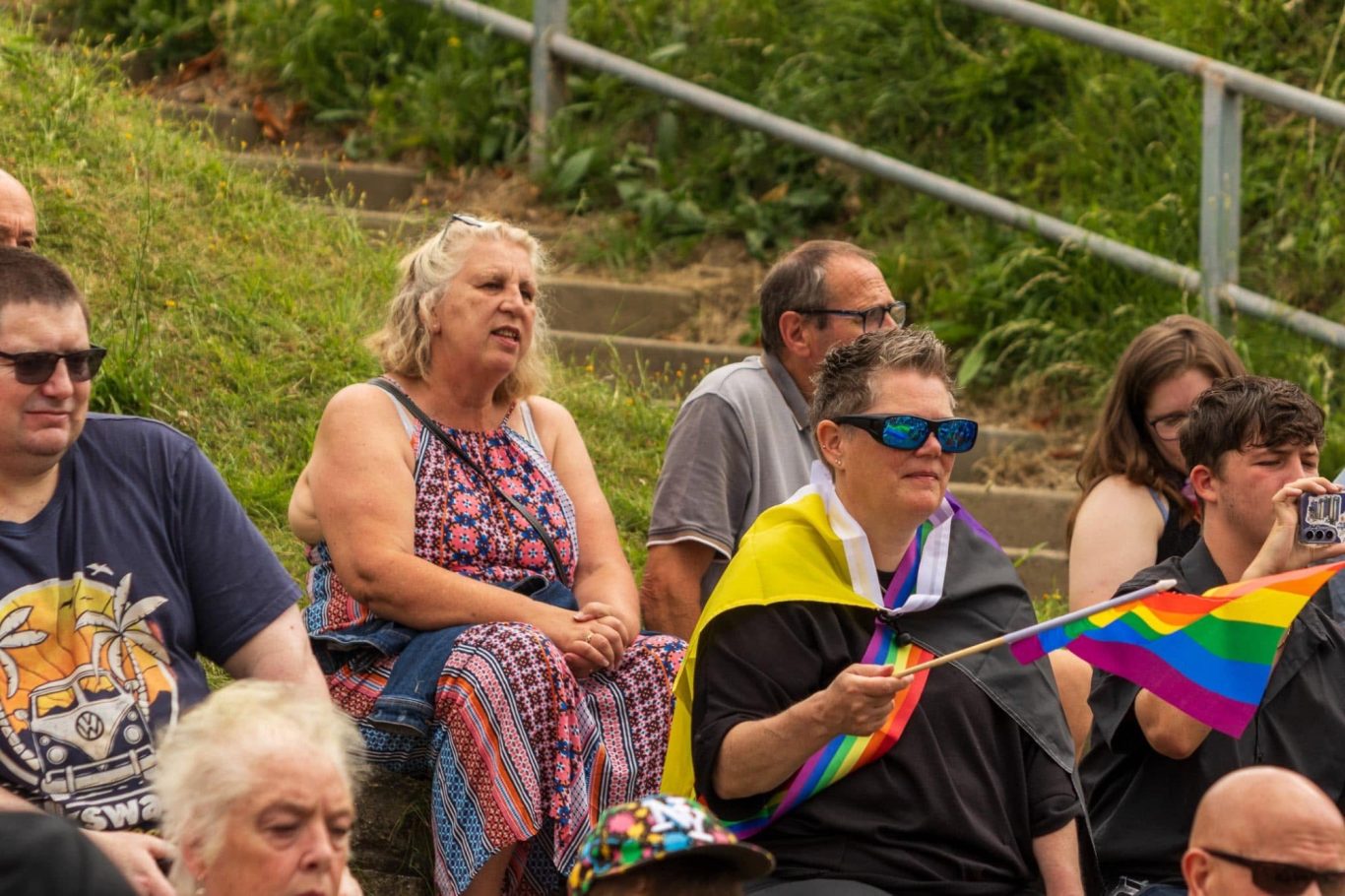 A diverse group of people seated outdoors, some holding a rainbow flag.