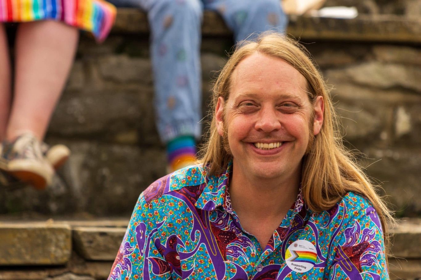 Smiling man with long hair wearing a colourful patterned shirt, seated on stone steps.