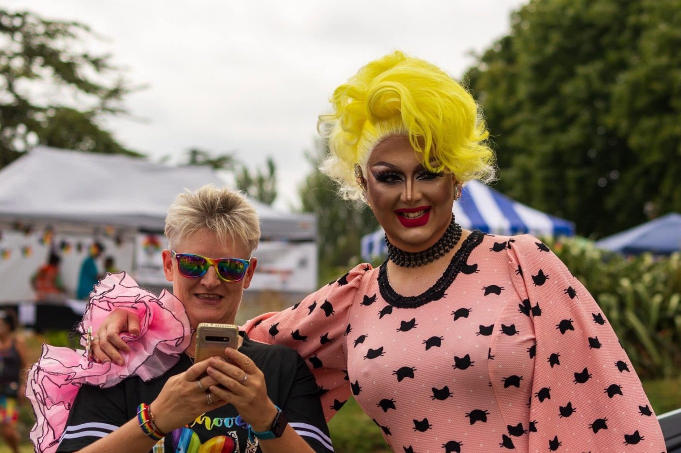Two people pose together outdoors; one in a pink, spotted outfit and bright yellow wig, the other wearing rainbow sunglasses.