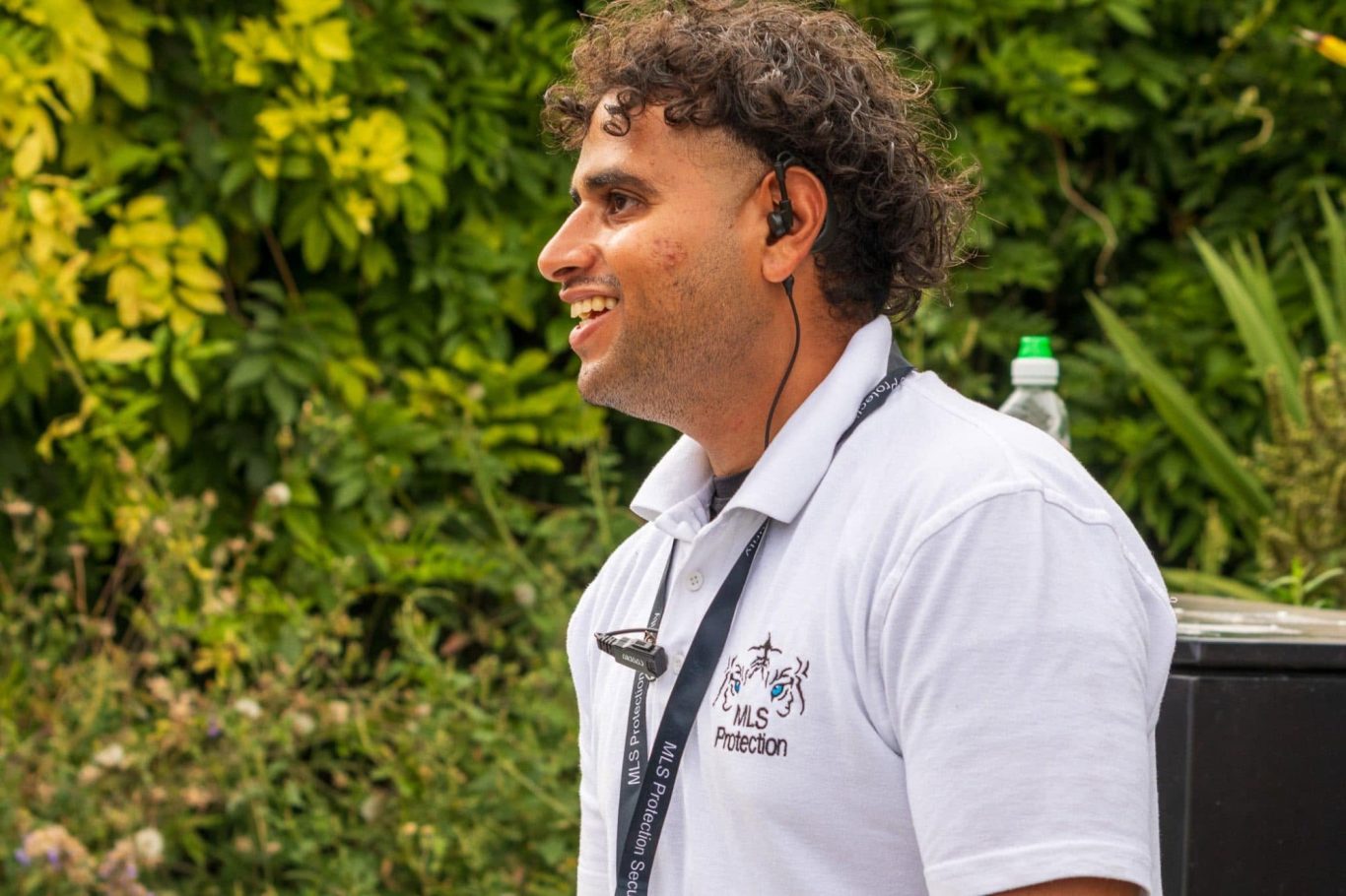 Smiling man in a white polo shirt with a headset, surrounded by greenery.