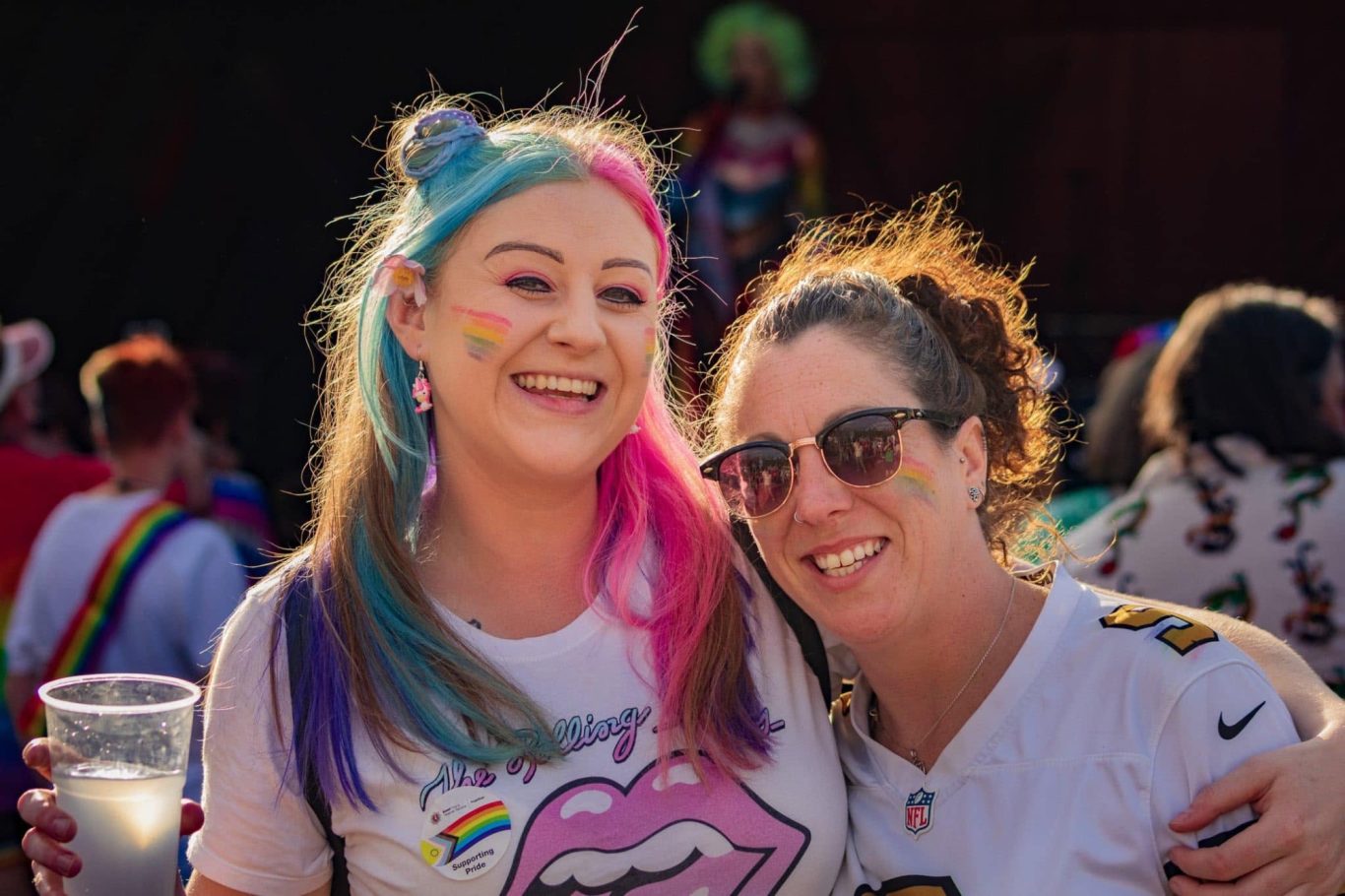 Two smiling women at a festive event, with rainbow-themed accessories and drinks.