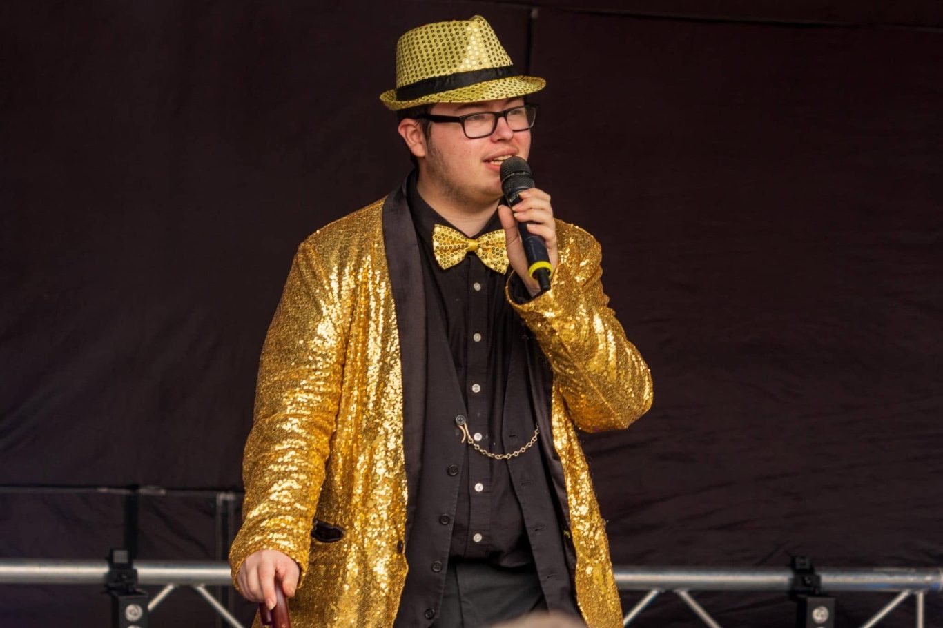GYG - 2024 A performer in a gold sequined jacket and hat, speaking into a microphone.