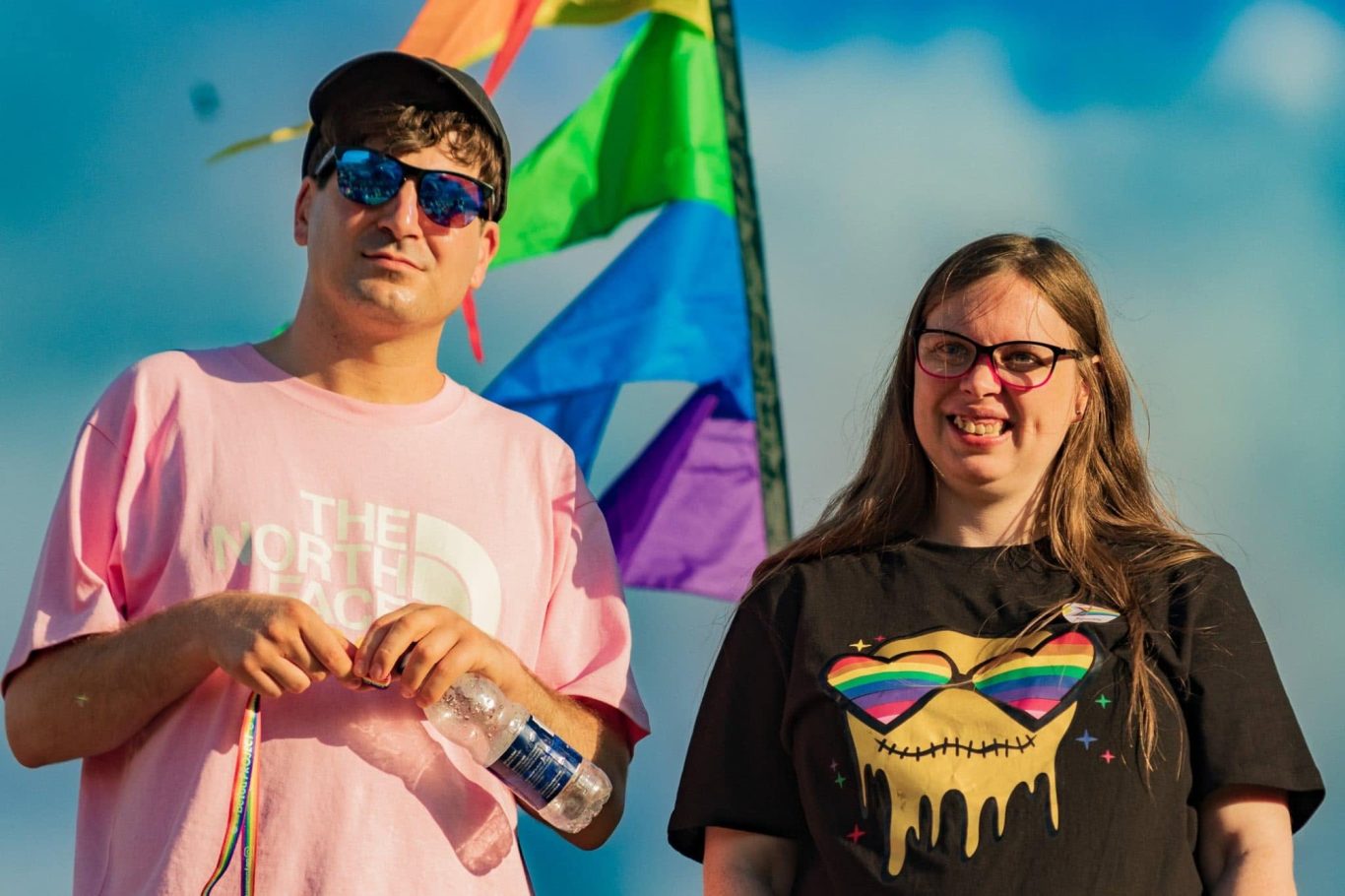 Two people stand together near colourful flags, smiling and wearing casual clothing.