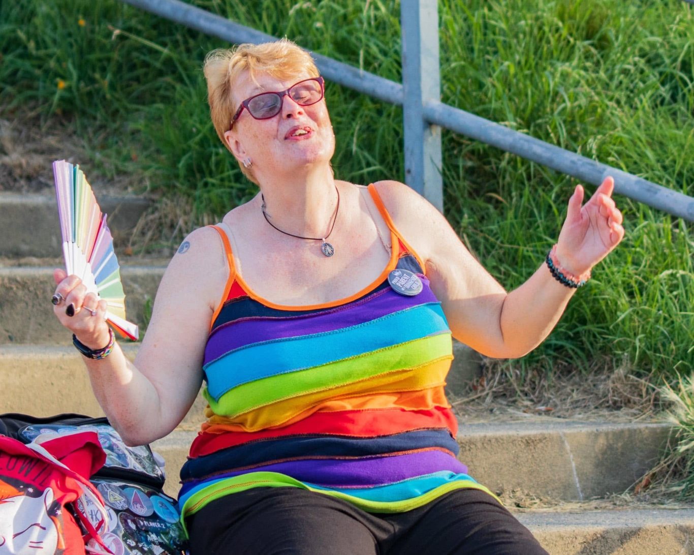 A woman in a rainbow tank top joyfully poses while sitting outdoors.