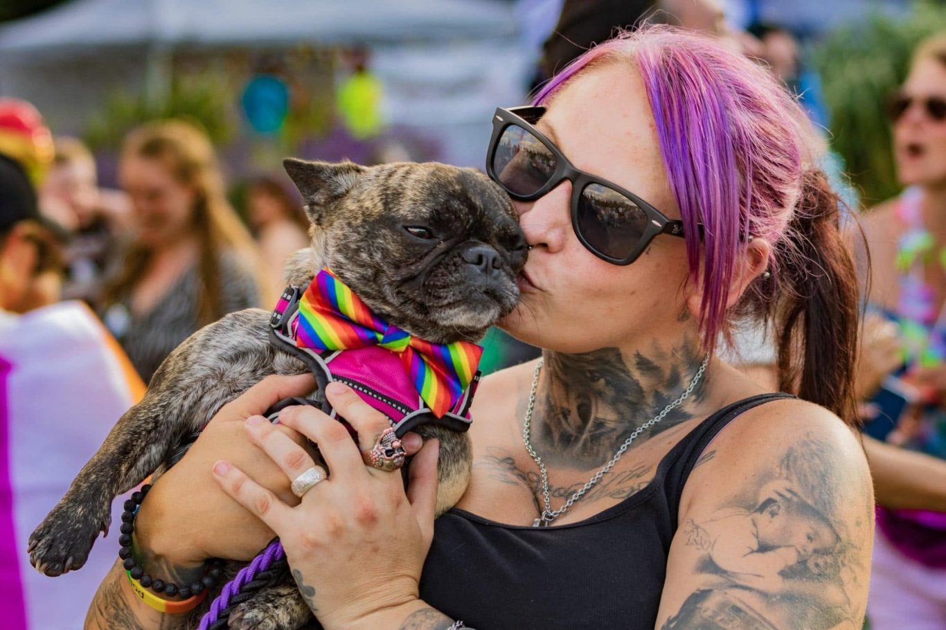A woman with sunglasses kisses a small dog wearing a rainbow scarf at a festive event.