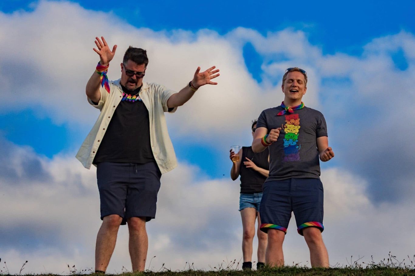 Two men celebrating outdoors, wearing rainbow accessories, with a person smiling in the background.
