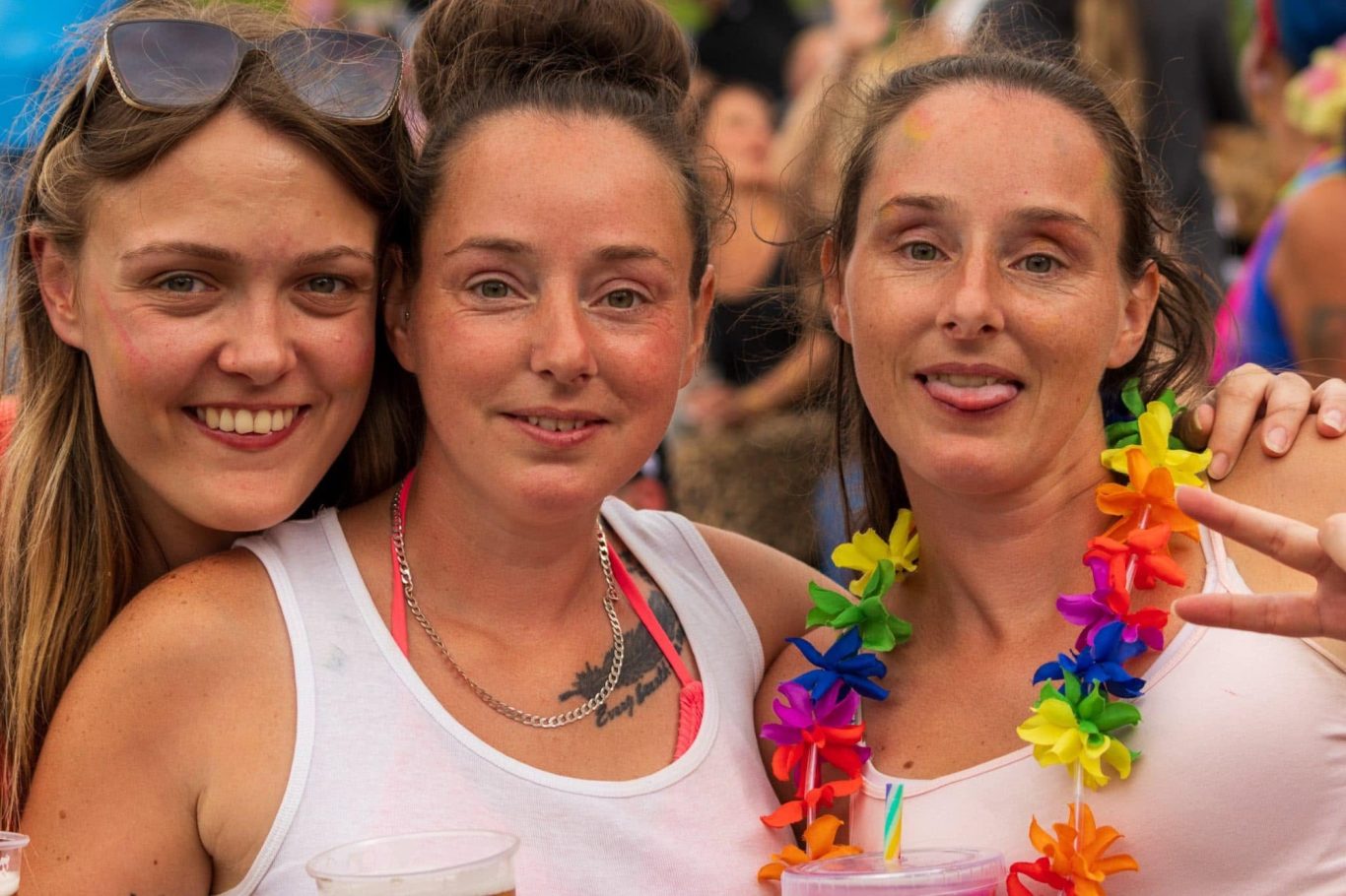 Three women smiling, wearing casual clothing and flower leis at a festive outdoor event.