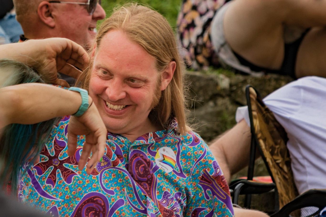 Man with long hair wearing a colourful shirt, smiling among a crowd.