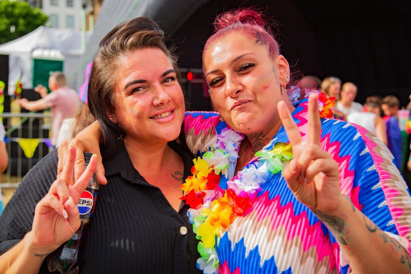 Two women smiling and posing together, wearing colourful leis at an outdoor event.