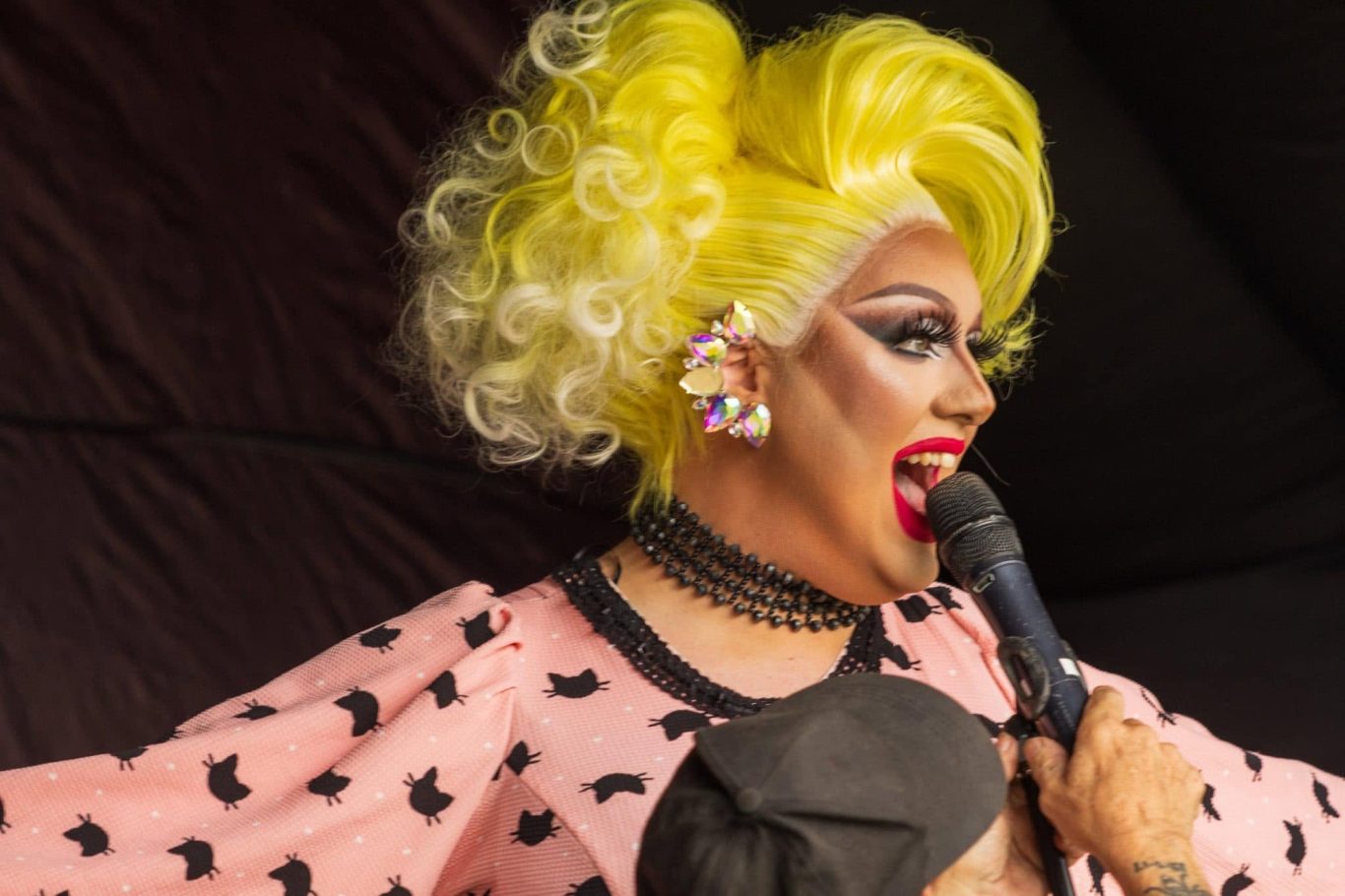 Amanda Bang - 2024 A drag performer with yellow curly hair and vibrant makeup holding a microphone.