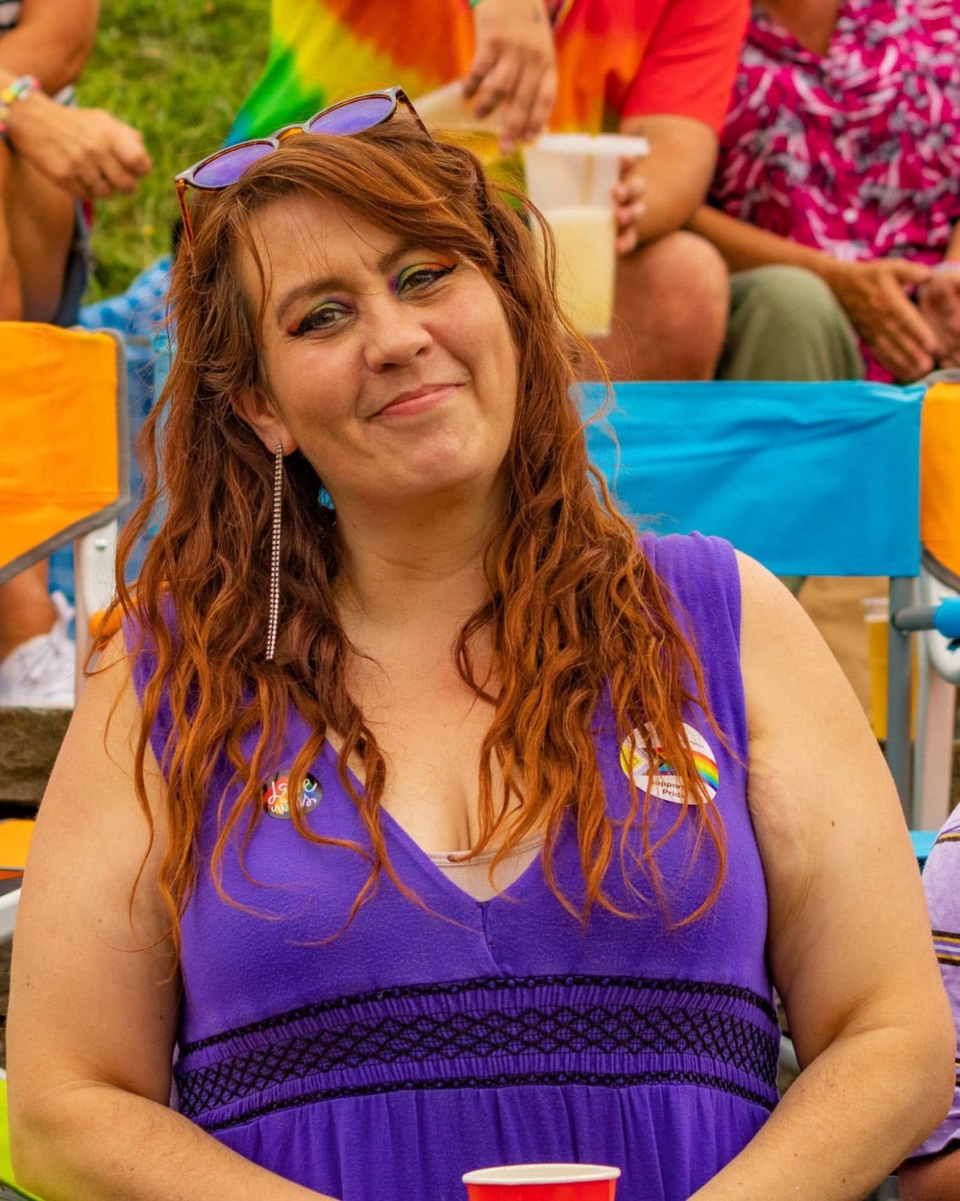 Smiling woman with long red hair, wearing a purple dress, holding a red cup.