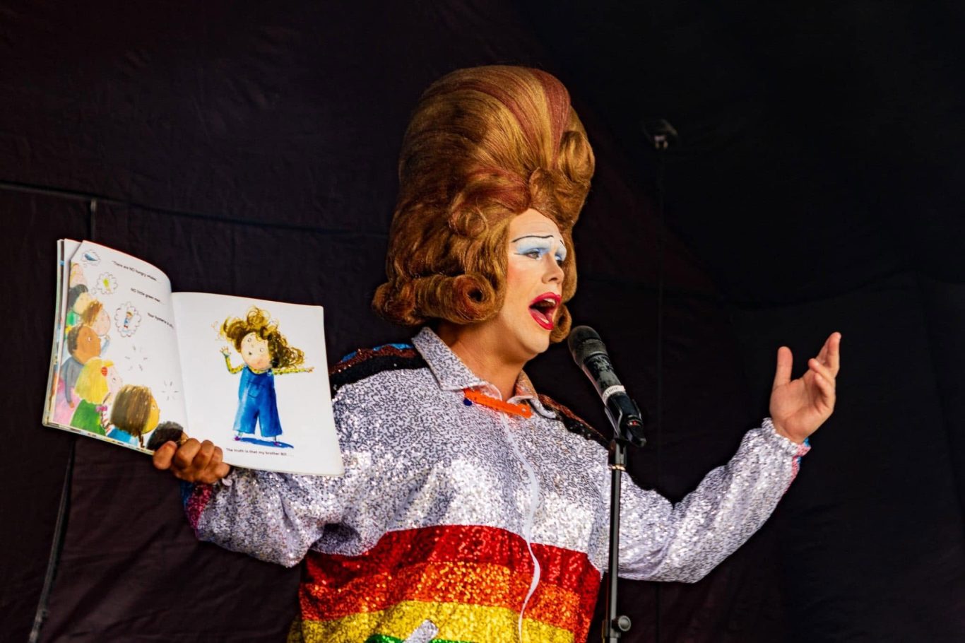 Mama G - 2024 A performer in drag reading a storybook on stage with theatrical expressions.