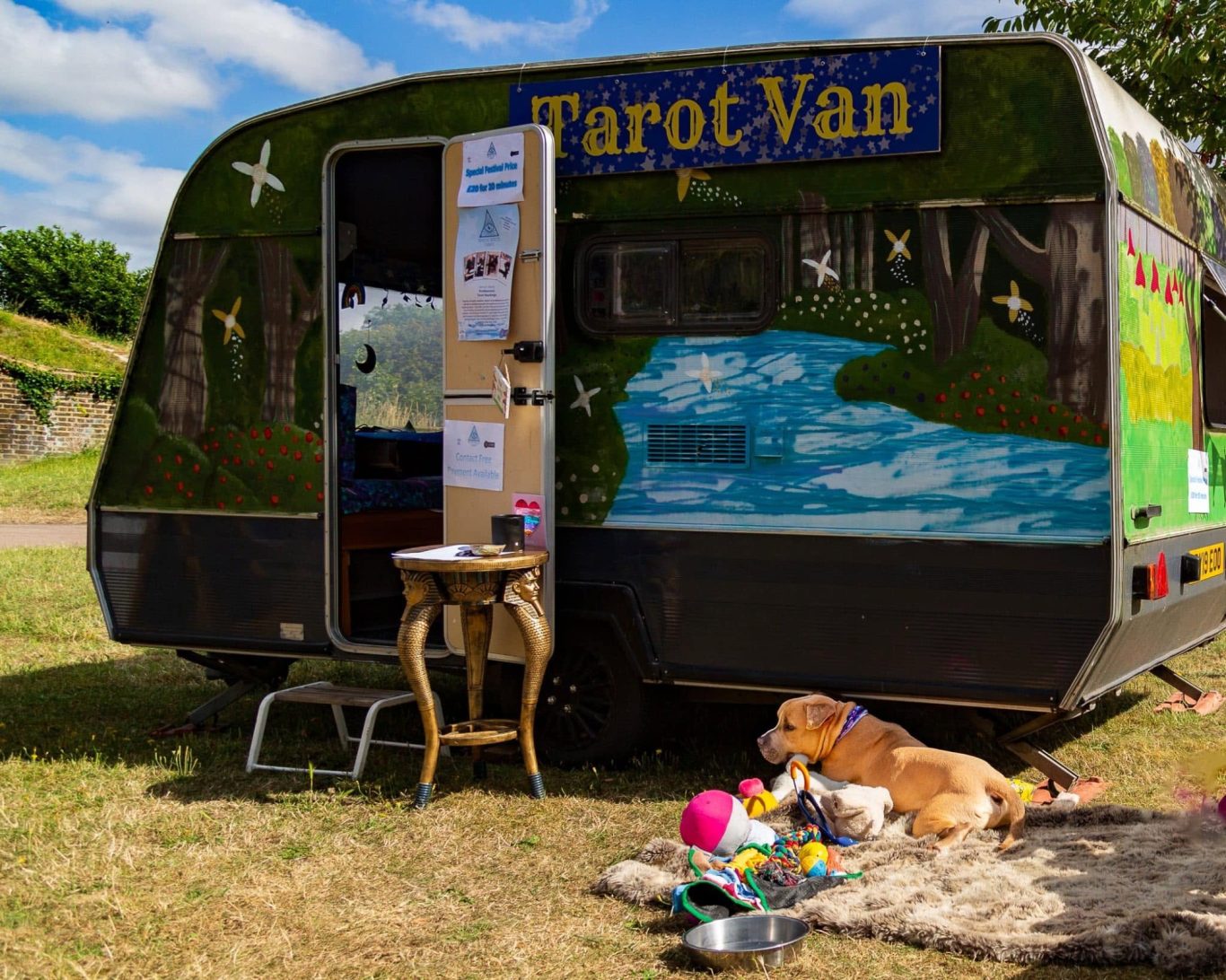 A colourful caravan for tarot reading, with a dog resting beside it on grass.