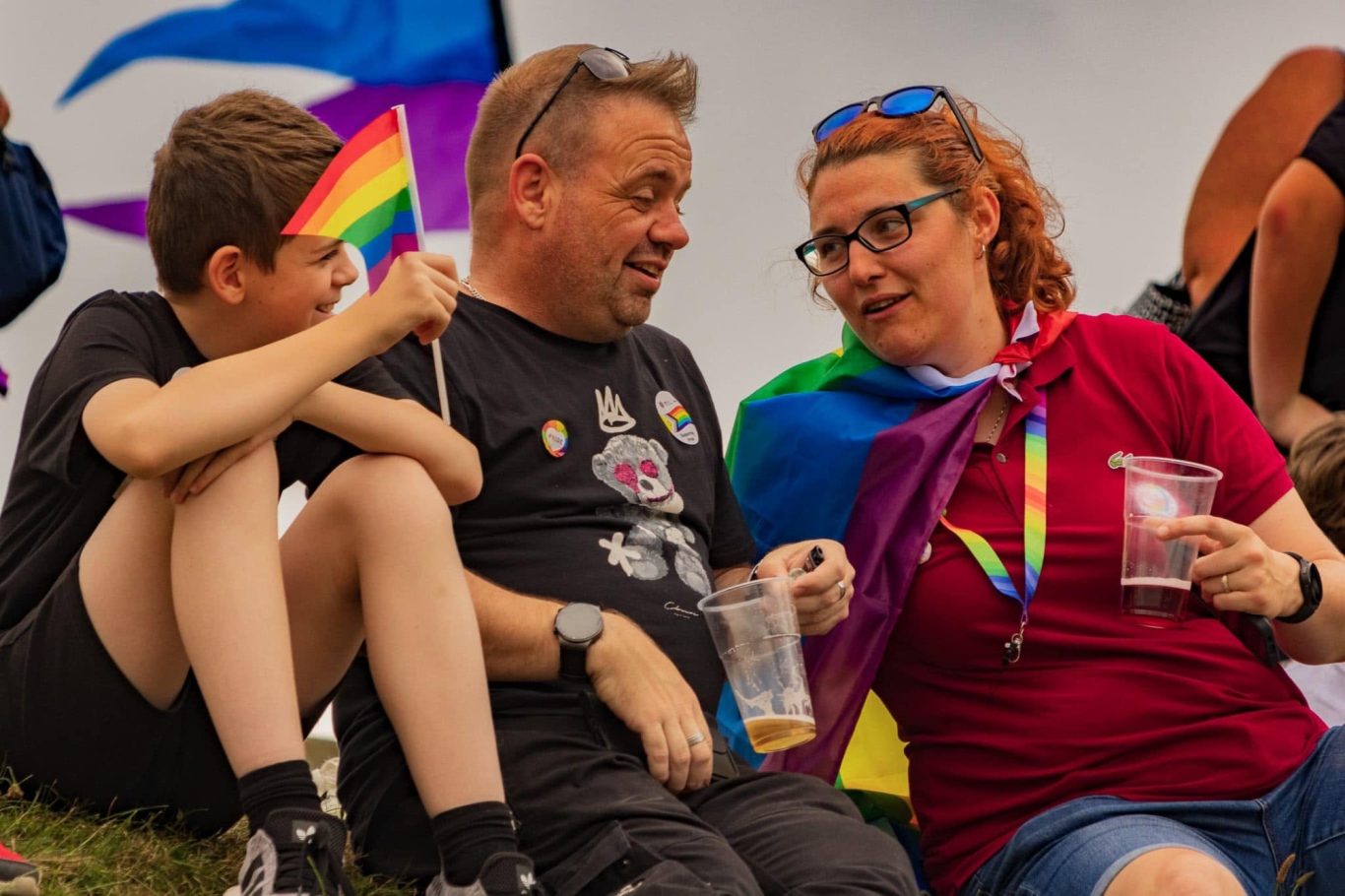 A family enjoying a pride event, holding flags and drinks, celebrating together.
