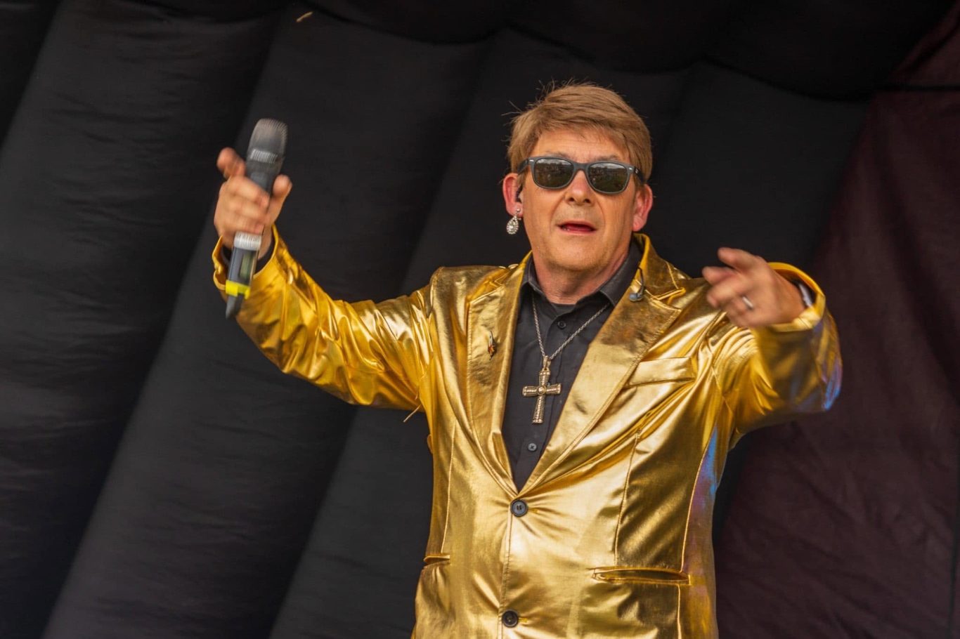 Elton John Tribute A performer in a gold jacket and sunglasses sings on stage.