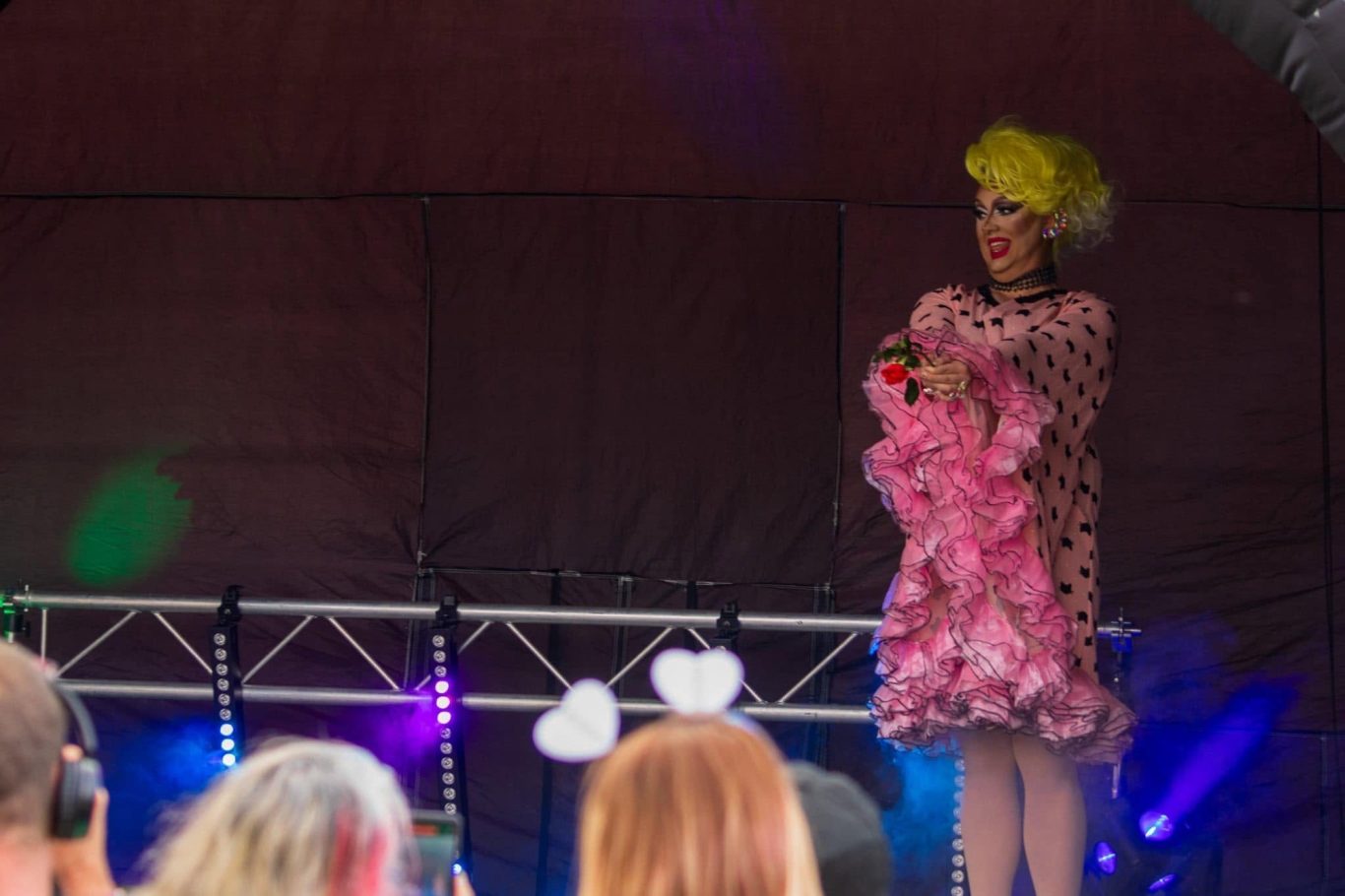 Marshall Arkley - 2024 A performer in a pink dress and yellow wig stands on stage, holding a pink feather boa.