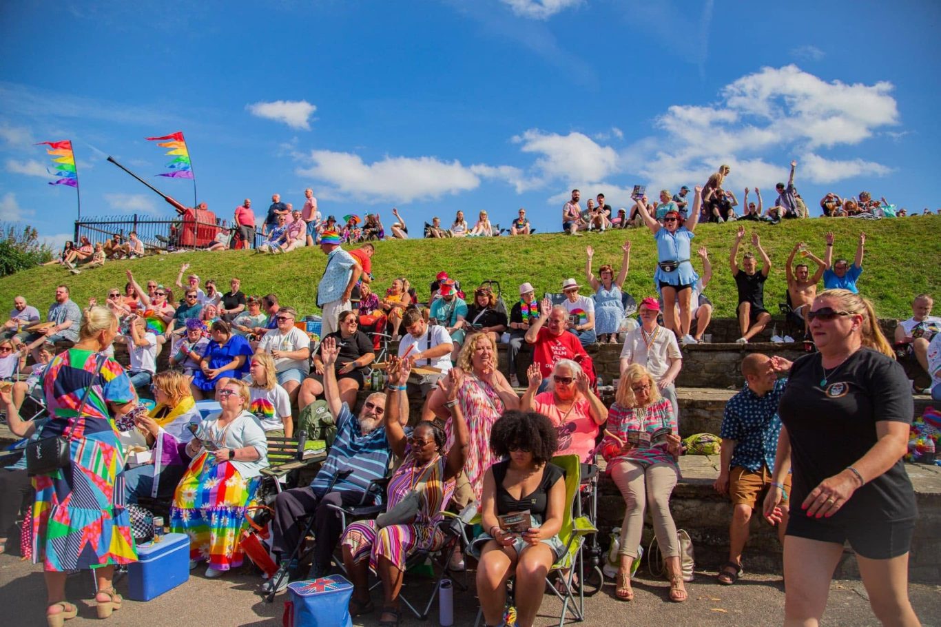 A vibrant crowd celebrating at an outdoor event with rainbow flags and diverse attendees.