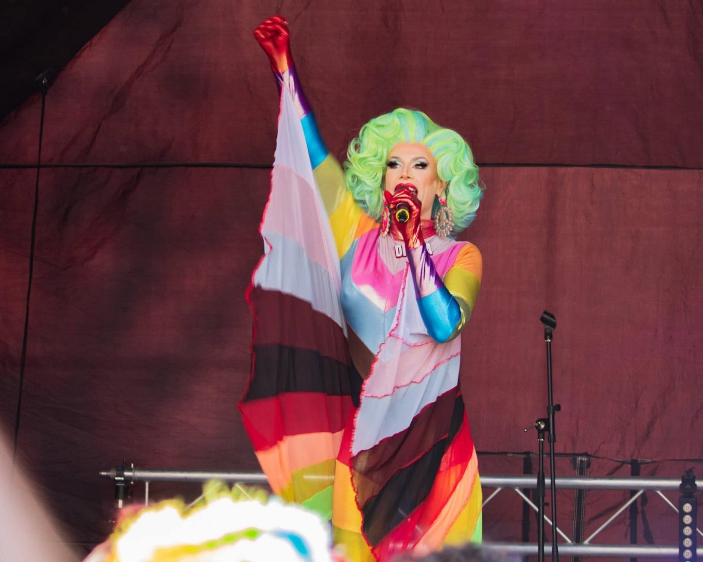 Divina DiCampo - 2024 A performer in vibrant costume with rainbow flags, singing passionately on stage.