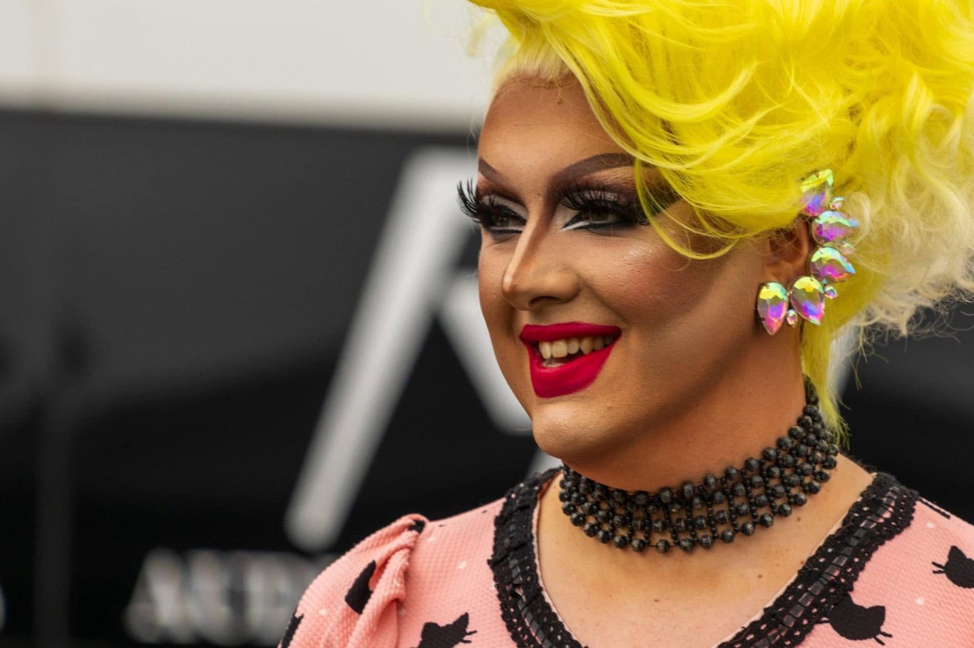 Person with bright yellow hair, colourful earrings, and bold makeup, smiling.