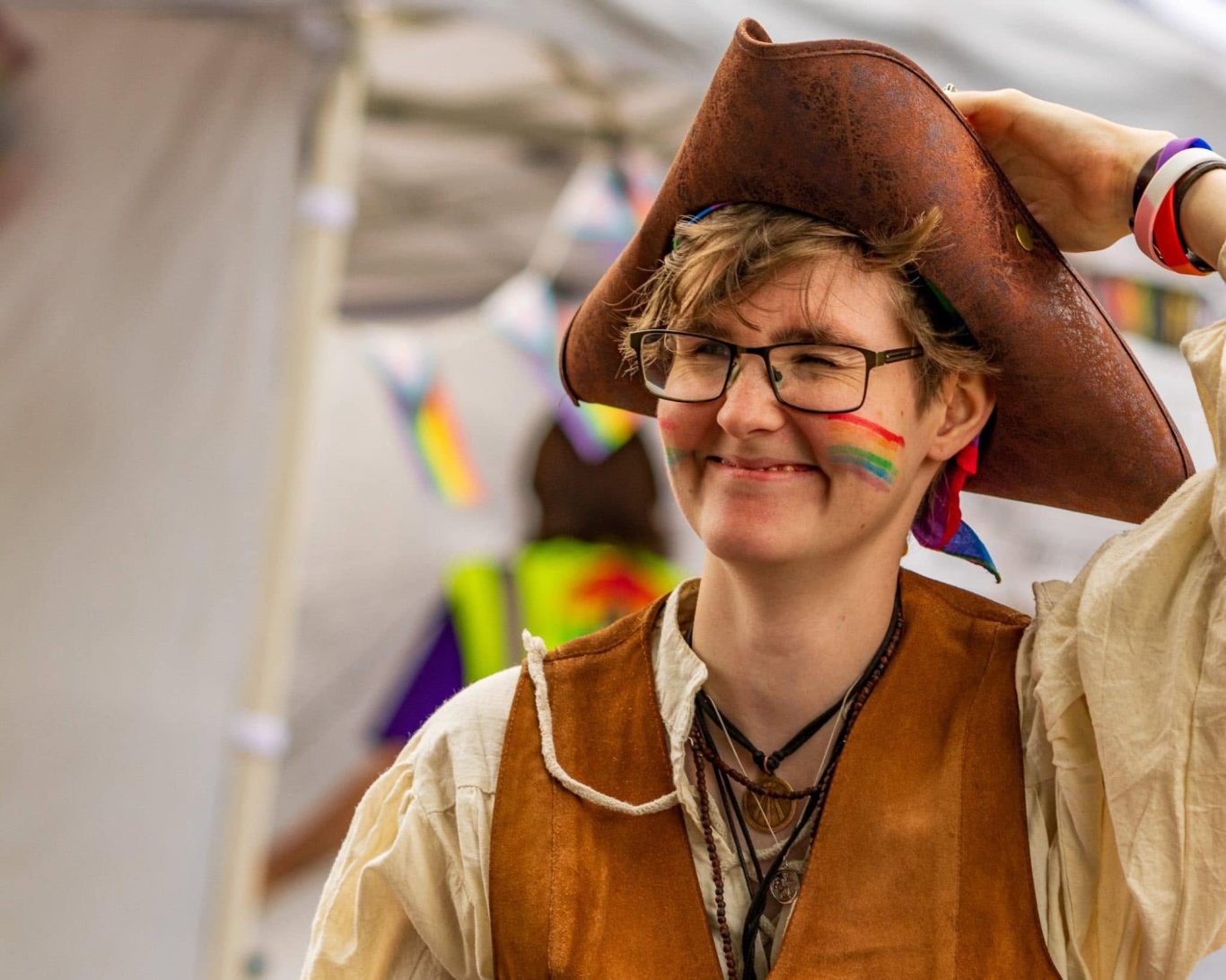 Smiling person wearing a pirate hat and rainbow face paint, in a festive setting.