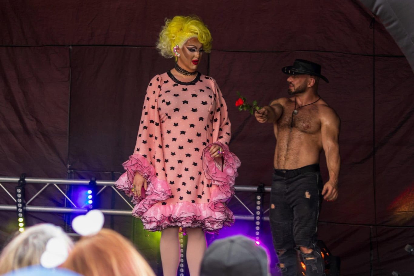 Marshall Arkley - 2024 Drag performer in pink polka dot dress with yellow wig, alongside a muscular man in a hat.