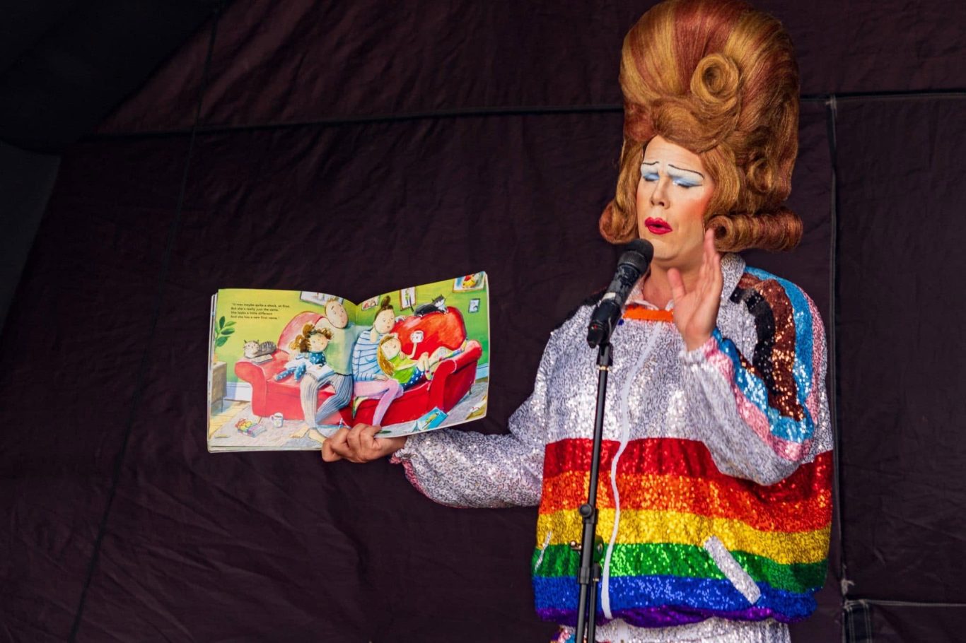 Mama G - 2024 A performer in a rainbow jacket reading a storybook on stage.