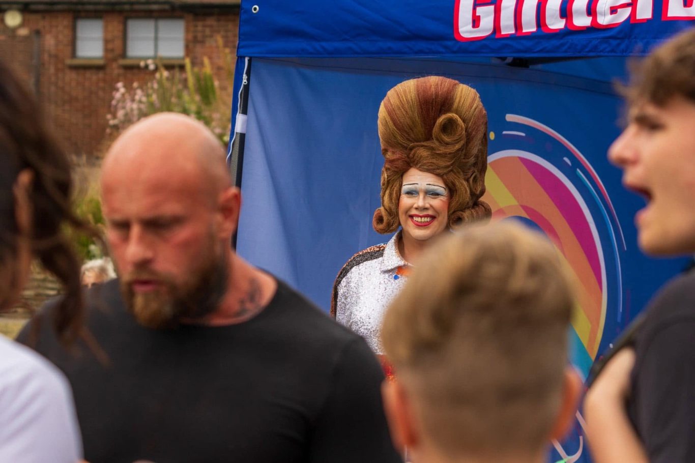 A person in a large wig smiles from behind a crowd at an outdoor event.