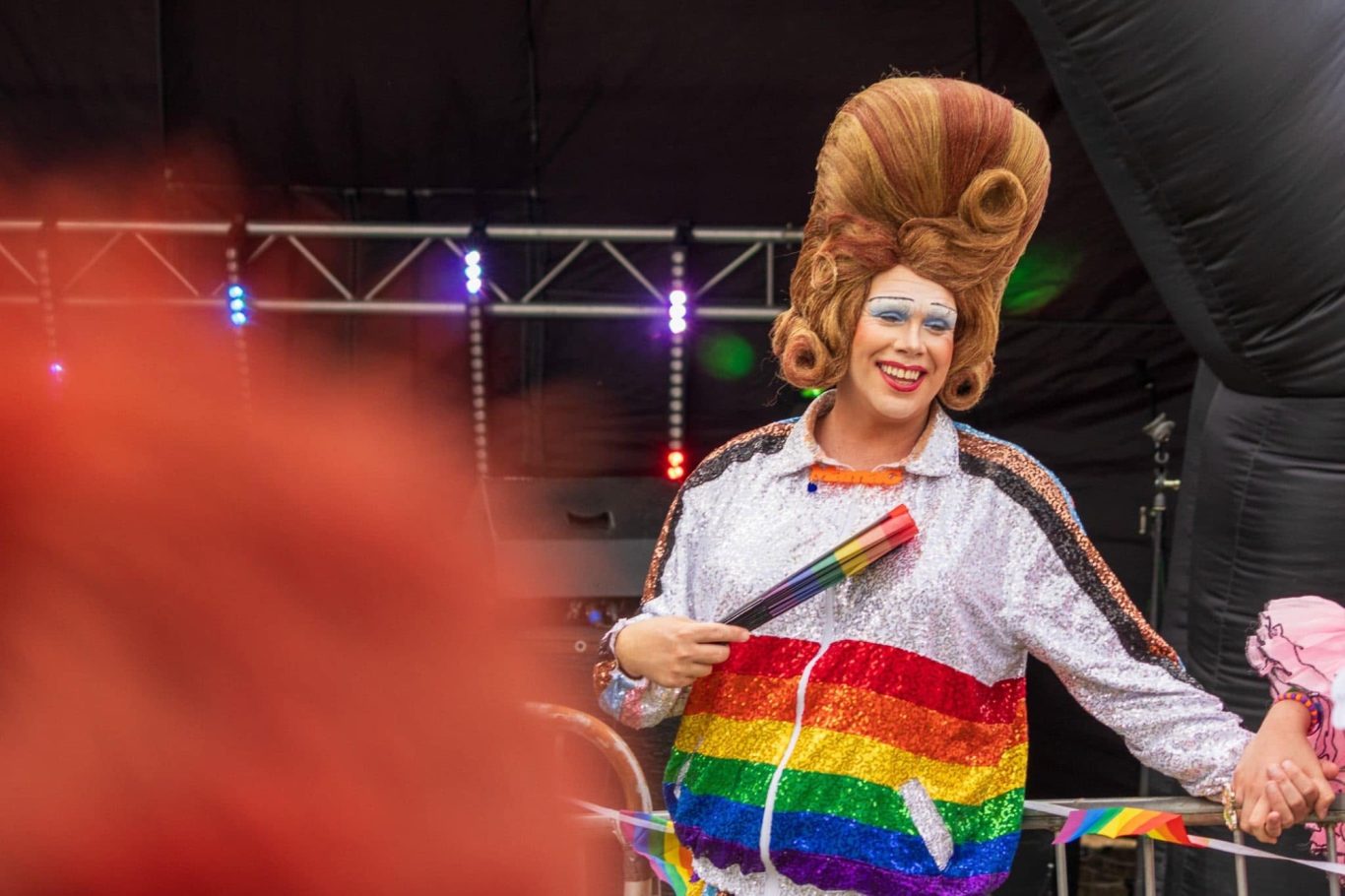 Mama G - 2024 Person in a large wig, wearing a rainbow-striped outfit, holding a rainbow flag.