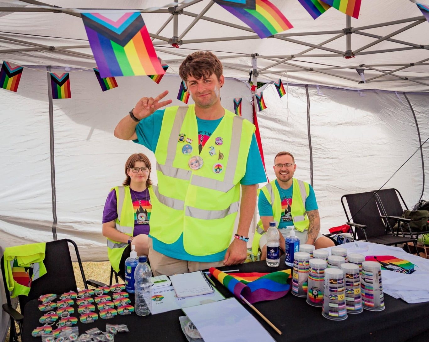 Three volunteers in bright vests at a celebration, surrounded by rainbow flags and drinks.