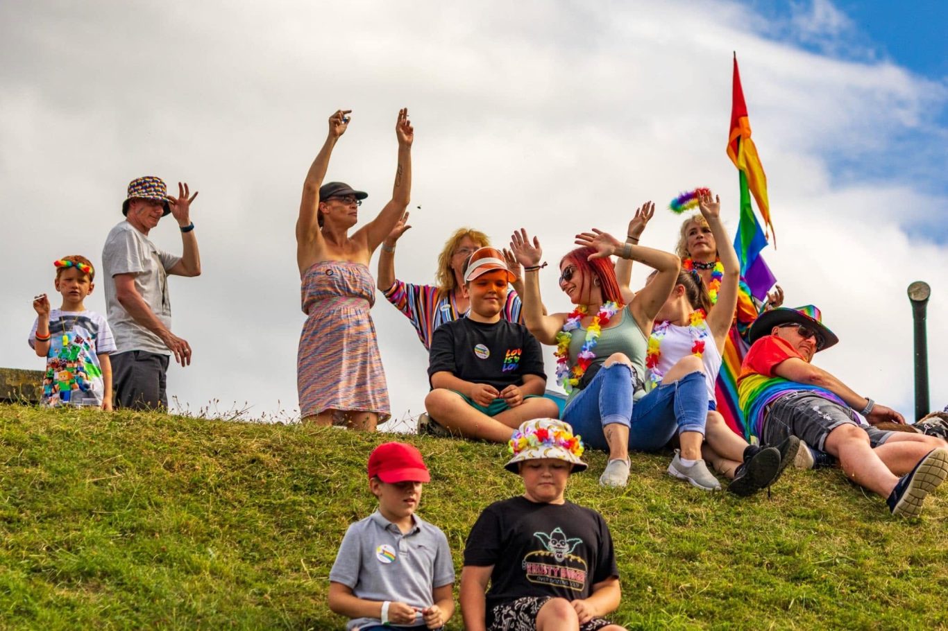 A diverse group of people celebrating with rainbow flags and festive outfits on a grassy hill.