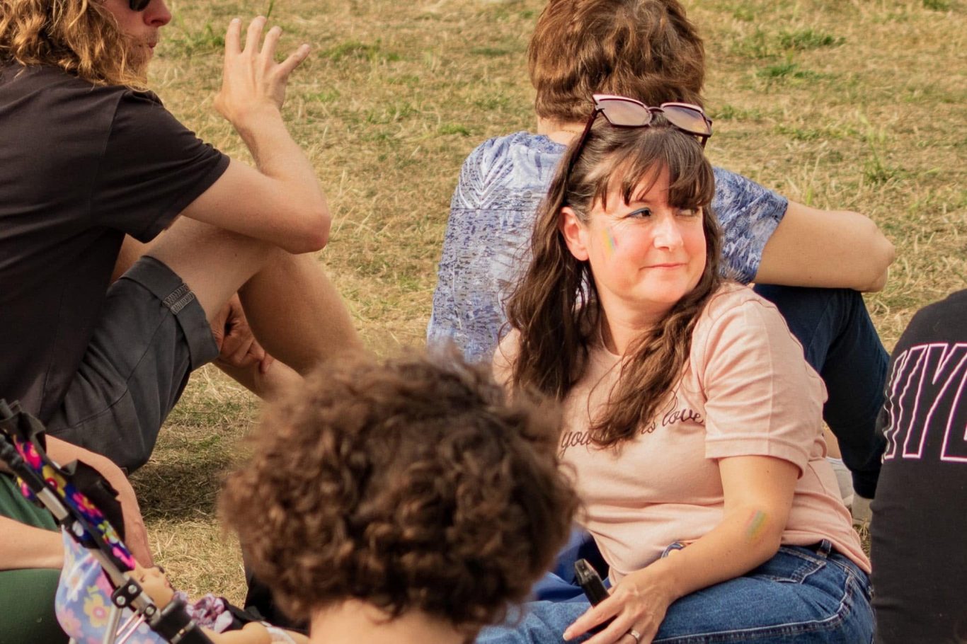 A woman with long hair and a light pink top sits on grass, looking thoughtfully.