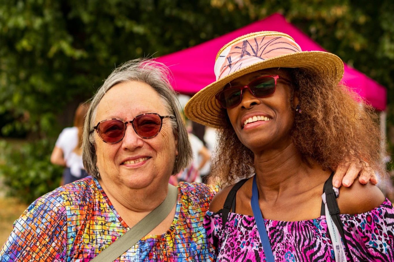 Two women smile together, wearing sunglasses and hats, with a pink tent in the background.