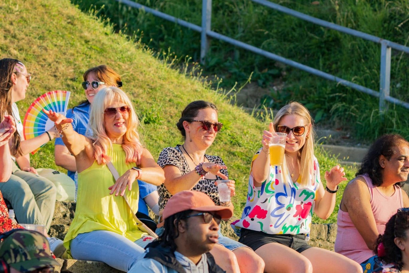 Three women in summer attire enjoying a sunny outdoor event, smiling and holding drinks.