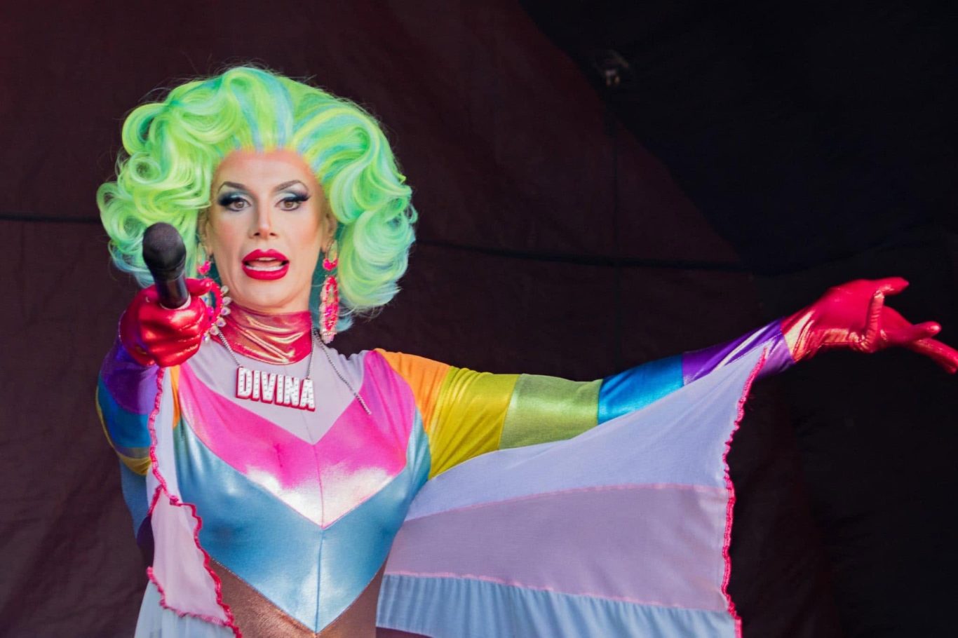 Divina DiCampo - 2024 A performer in vibrant drag attire with green hair, gesturing and holding a microphone.