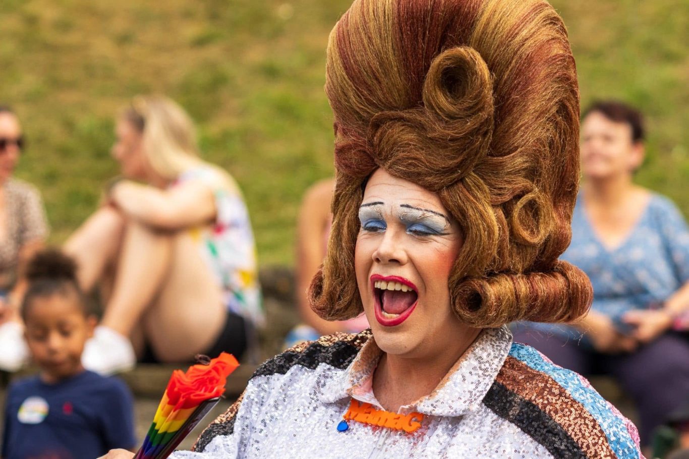 A performer in extravagant drag makeup and hair celebrates at a festive outdoor event.