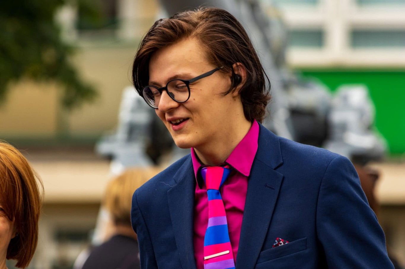 A young man in a blue suit and pink tie smiles, wearing glasses, outdoors.