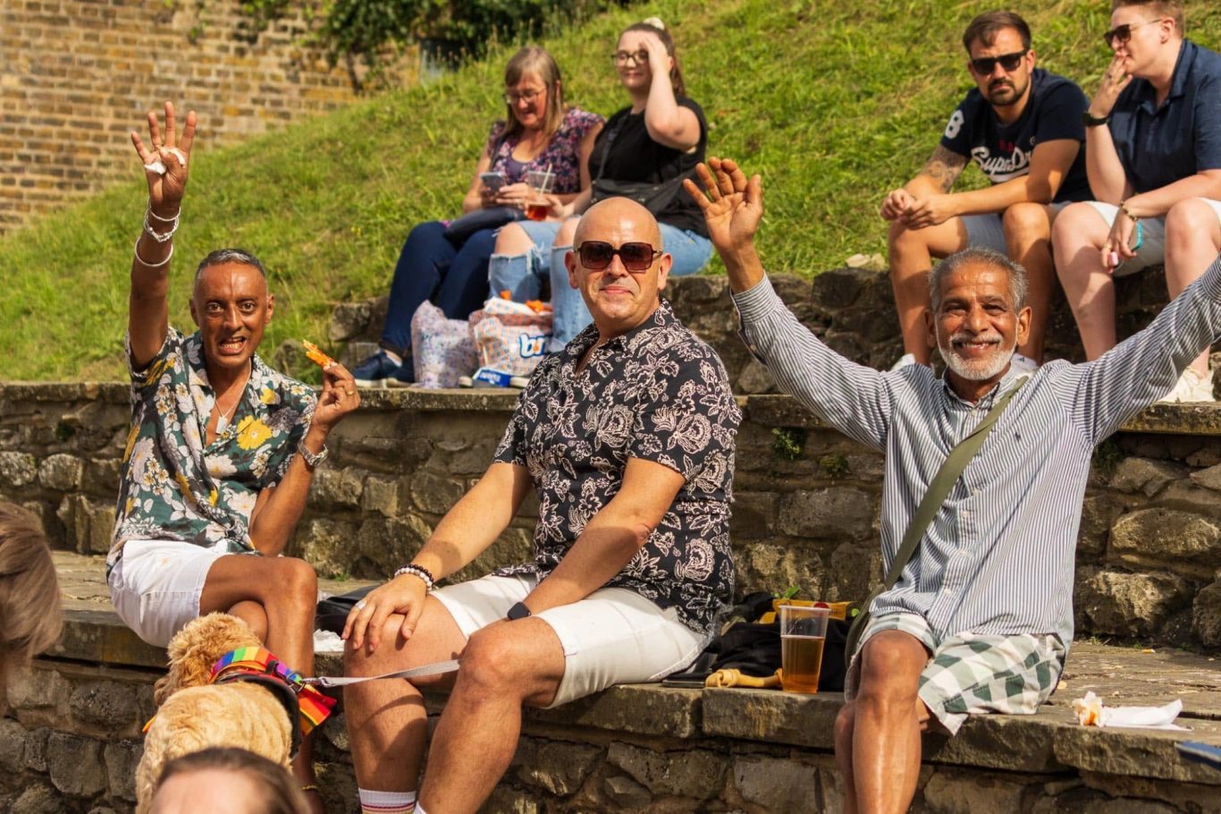 Three men sitting on a stone wall, waving and smiling, with a lively crowd in the background.