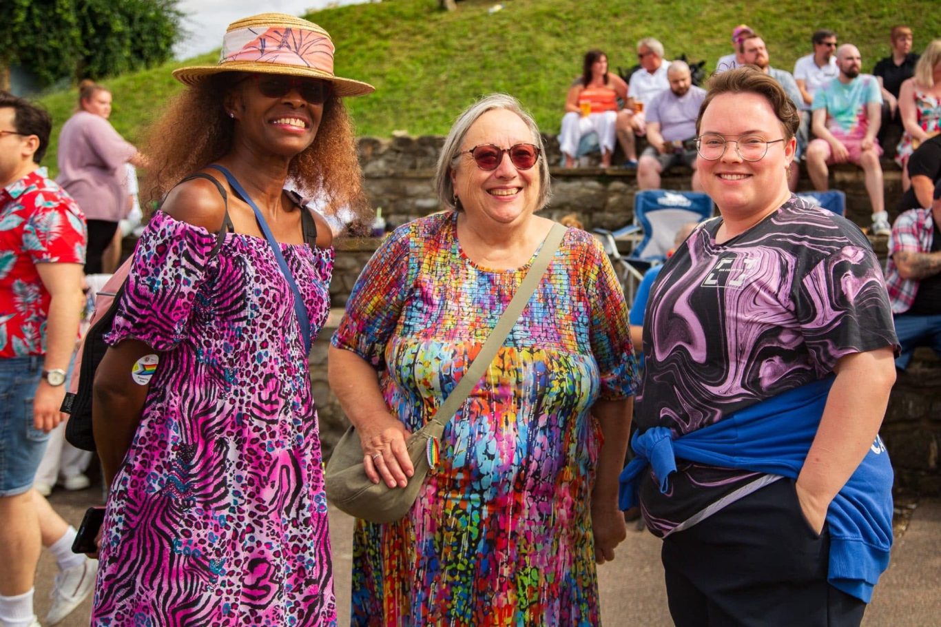Three women smiling together, dressed in colourful outfits at an outdoor event.