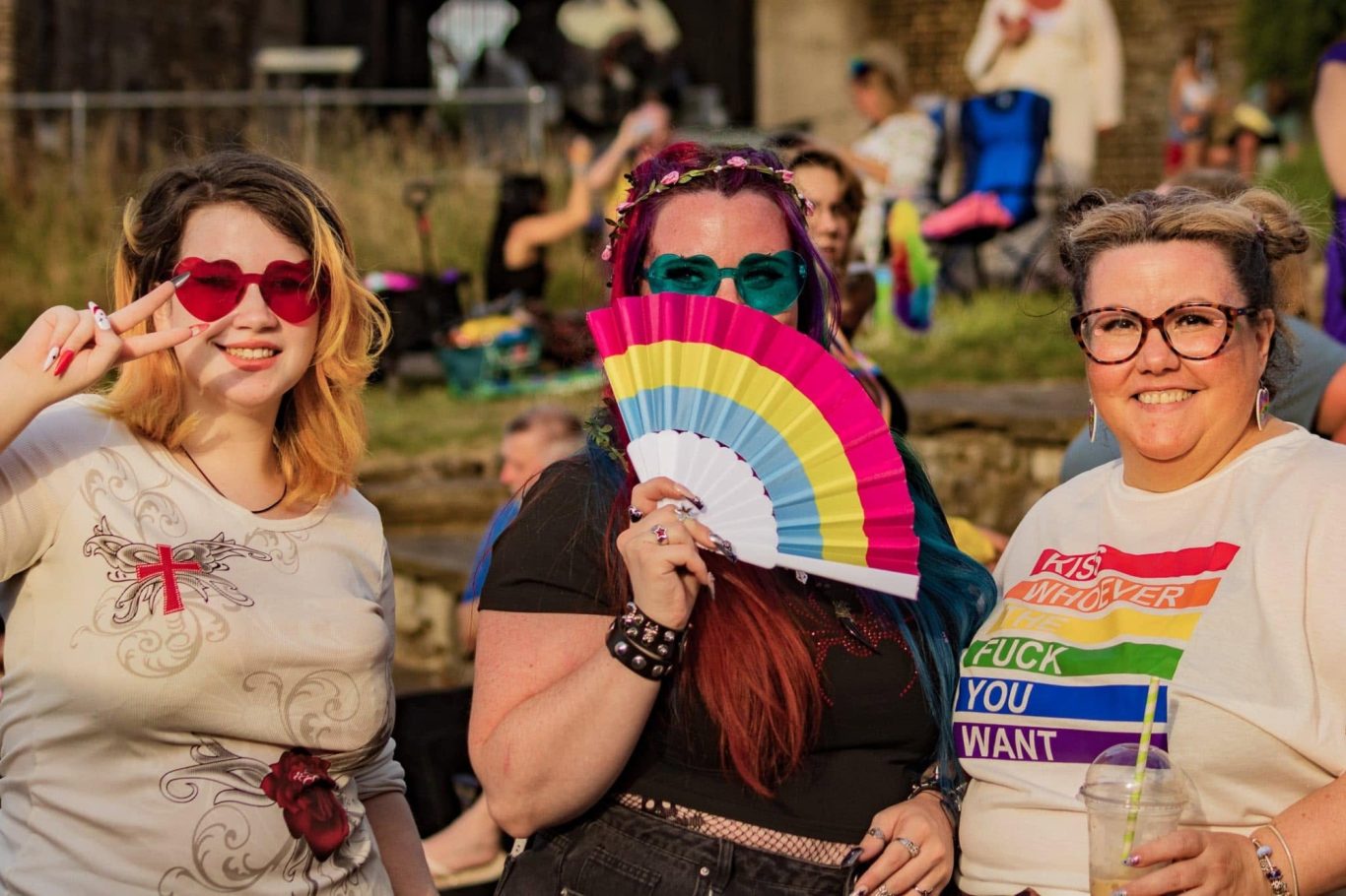 Three individuals at a celebration, one holding a rainbow fan, wearing vibrant accessories.